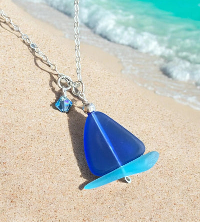 Handmade sailboat necklace with blue sea glass pendant and sterling silver chain on sandy beach background