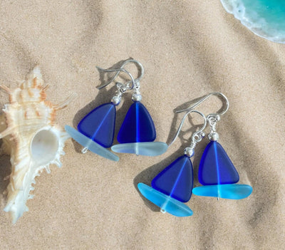 Sailboat sea glass earrings in cobalt blue with sterling silver wire wrapping on sandy beach background