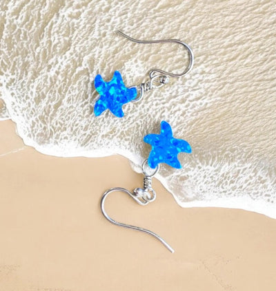 Sterling silver starfish earrings with blue opal charms on sandy beach background