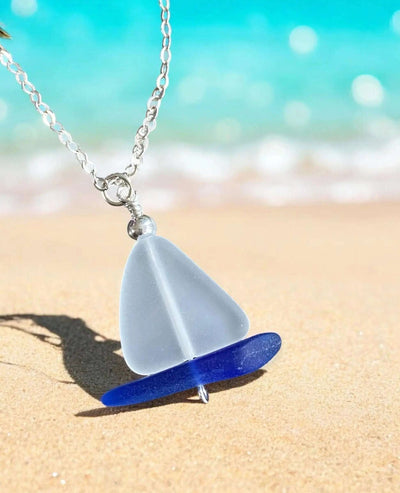 Sterling silver sailboat sea glass necklace with frosted white and blue glass pendant on beach background